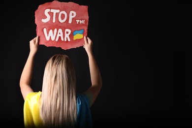 Photo of Woman wrapped in Ukrainian flag and holding poster with words Stop the War on black background, back view. Space for text