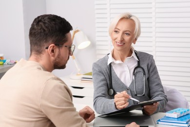 Photo of Doctor with pen and clipboard consulting patient at table in clinic