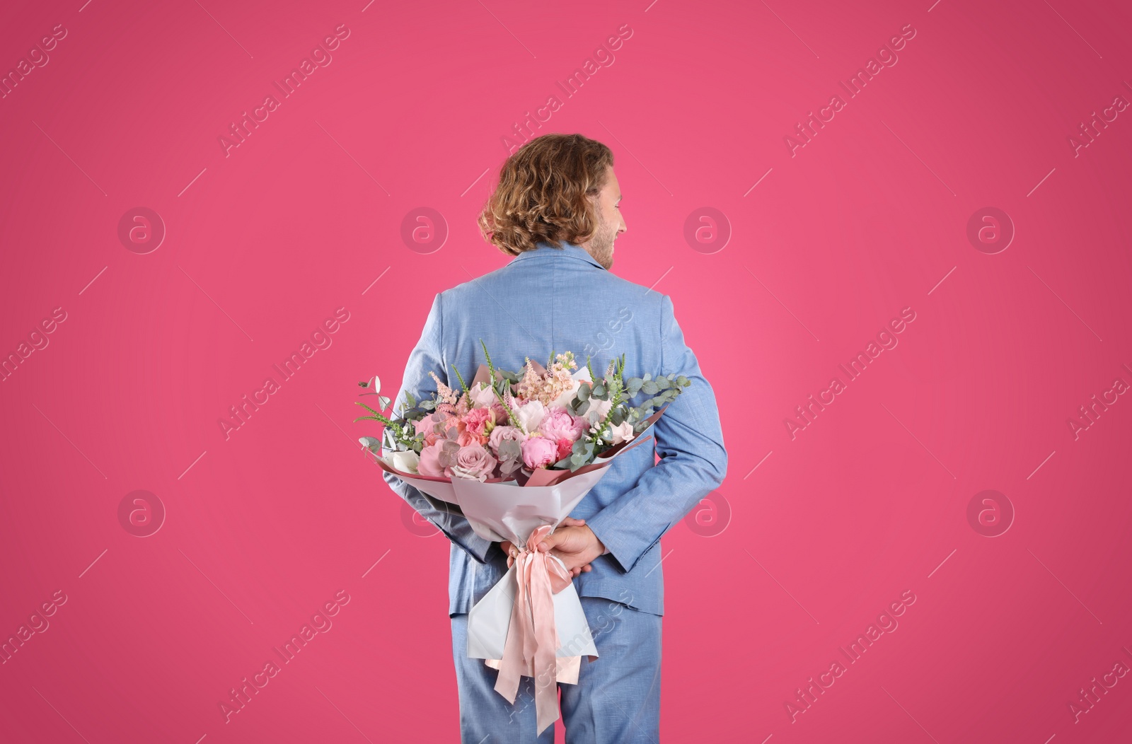 Photo of Man in stylish suit hiding beautiful flower bouquet behind his back on pink background