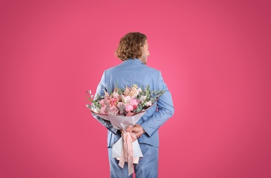 Photo of Man in stylish suit hiding beautiful flower bouquet behind his back on pink background