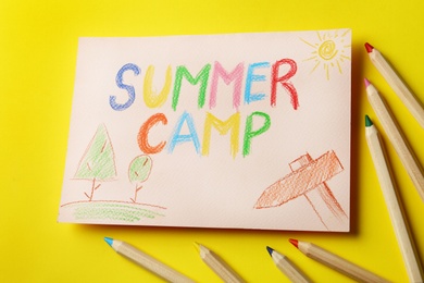 Paper with written text SUMMER CAMP, drawings and different pencils on color background, view from above