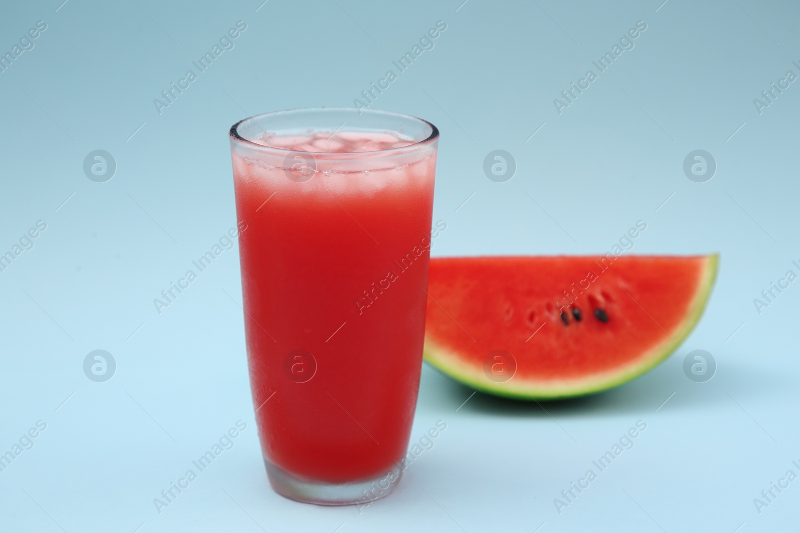 Photo of Glass of delicious drink with ice cubes and cut fresh watermelon on light blue background