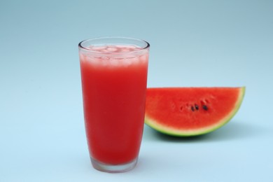 Glass of delicious drink with ice cubes and cut fresh watermelon on light blue background