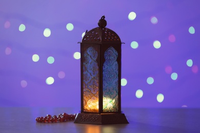 Photo of Muslim lamp Fanus with prayer beads on blurred lights background