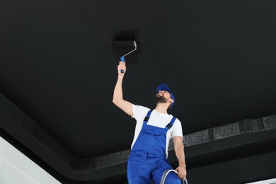 Worker in uniform painting ceiling with roller indoors