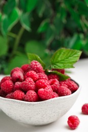 Photo of Bowl of fresh ripe raspberries with green leaves on white table against blurred background, closeup. Space for text