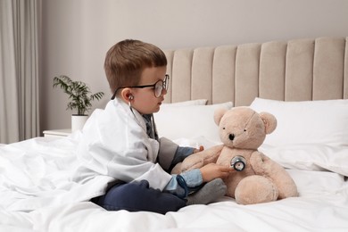 Cute little boy in pediatrician's uniform playing with stethoscope and toy bear at home