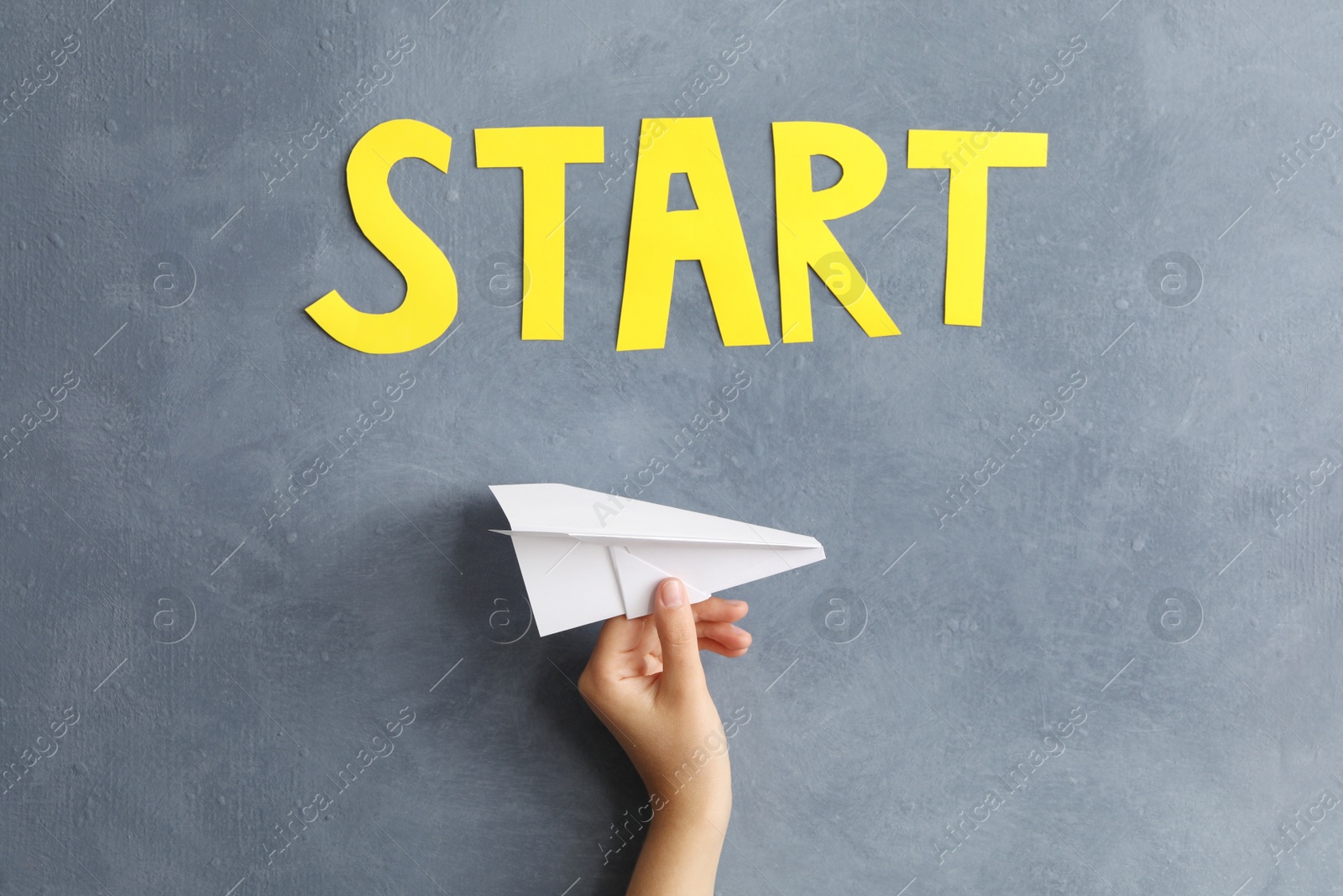 Photo of Woman holding paper plane near grey wall with word Start, closeup