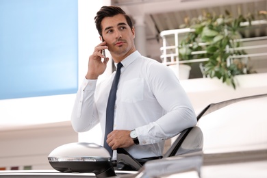 Photo of Young man talking on phone near car in modern dealership