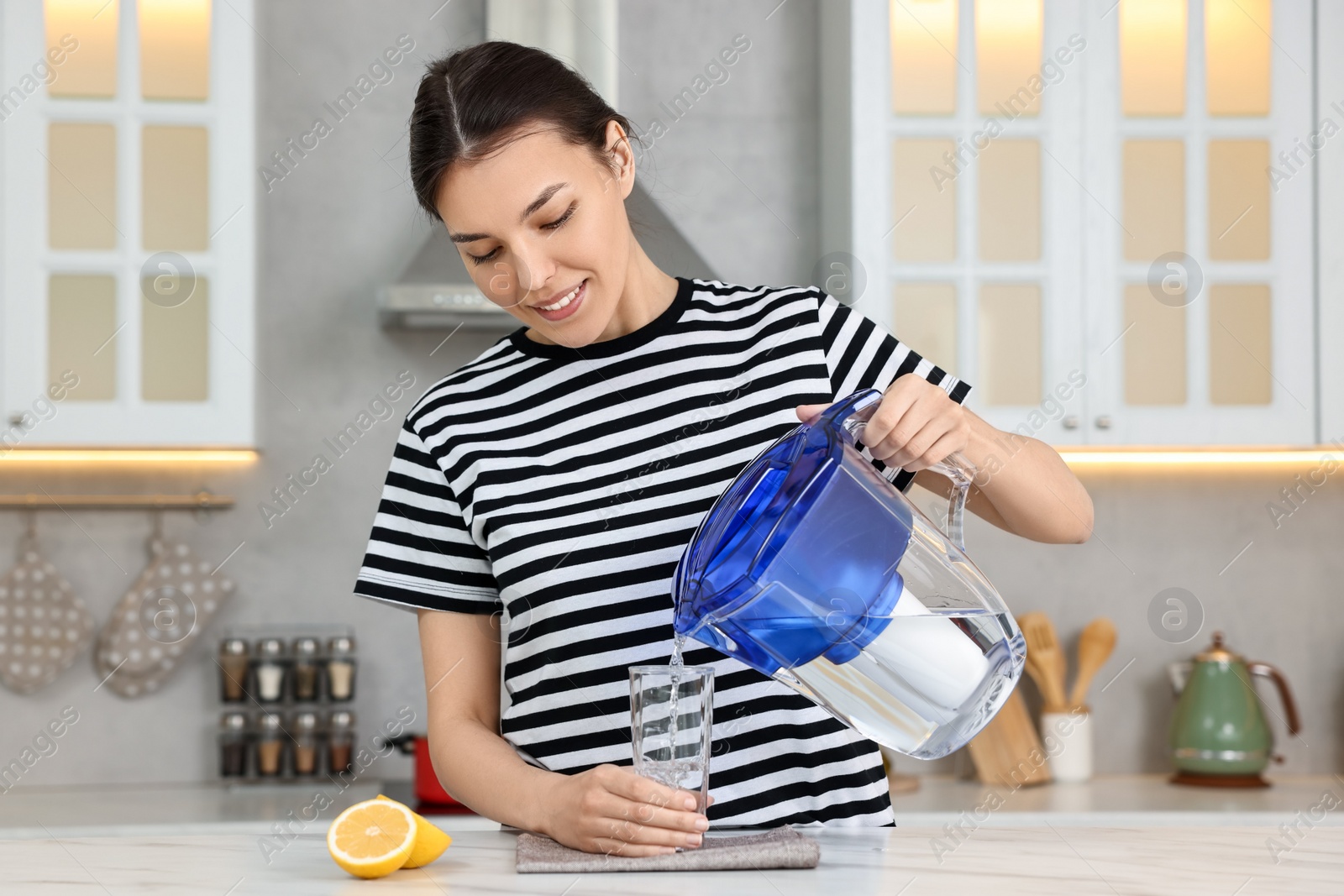 Photo of Woman pouring water from filter jug into glass in kitchen