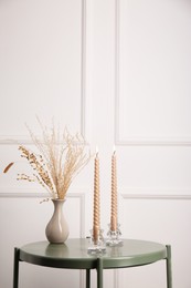 Dry plants in vase and burning candles on table near white wall