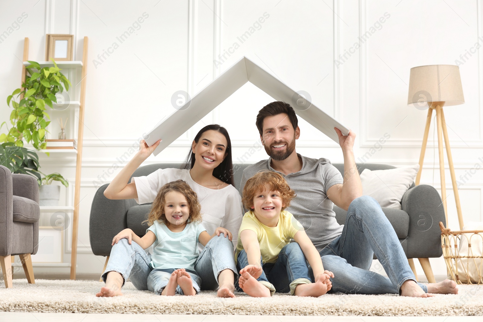 Photo of Family housing concept. Happy woman and her husband holding plastic roof while sitting with kids on floor at home