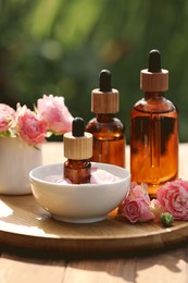 Bottles of rose essential oil and flowers on wooden table outdoors, closeup