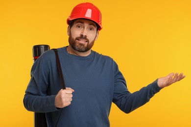 Photo of Confused architect in hard hat with drawing tube on orange background