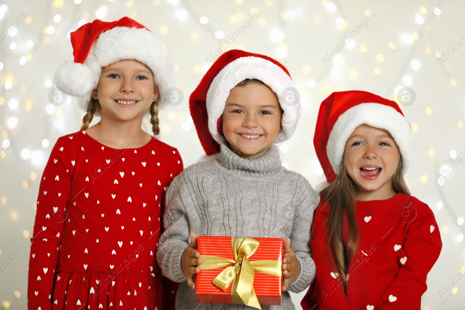 Photo of Happy little children in Santa hats with gift box against blurred festive lights. Christmas celebration