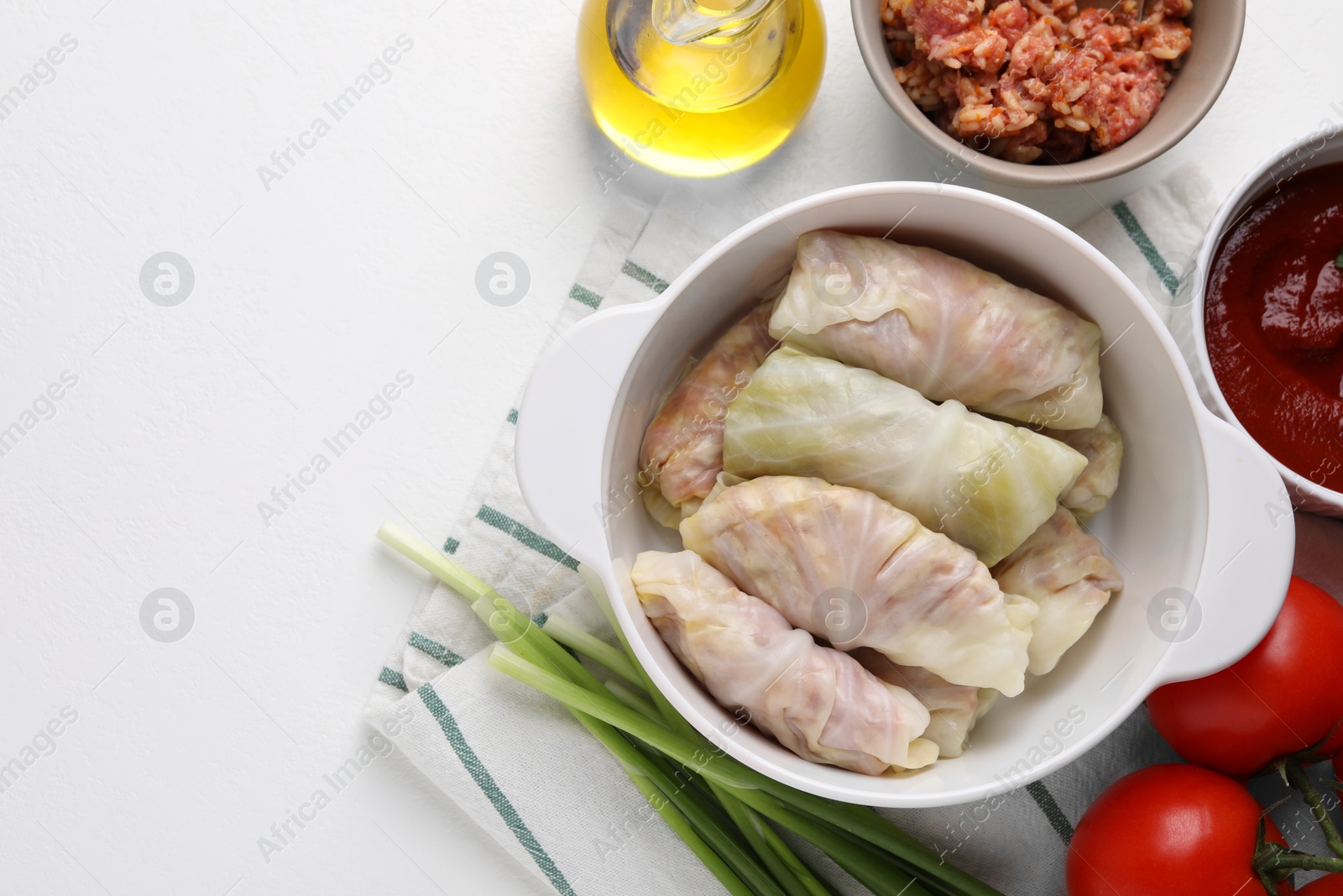 Photo of Ceramic pot with uncooked stuffed cabbage rolls and ingredients on white table, flat lay. Space for text