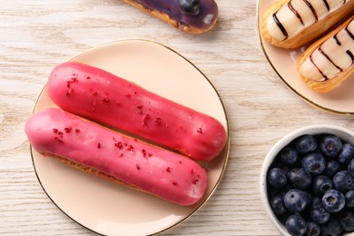 Photo of Different tasty glazed eclairs and blueberries on light wooden table, flat lay