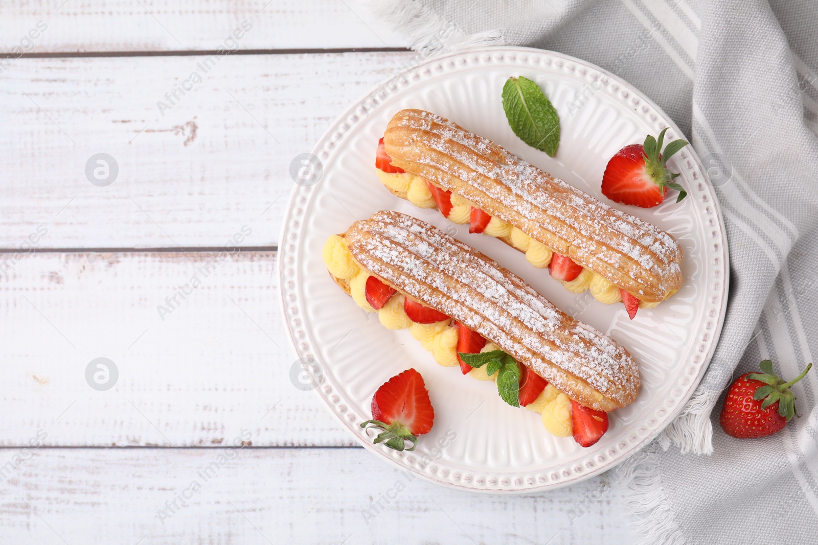 Photo of Delicious eclairs filled with cream, strawberries and mint on white wooden table, top view. Space for text