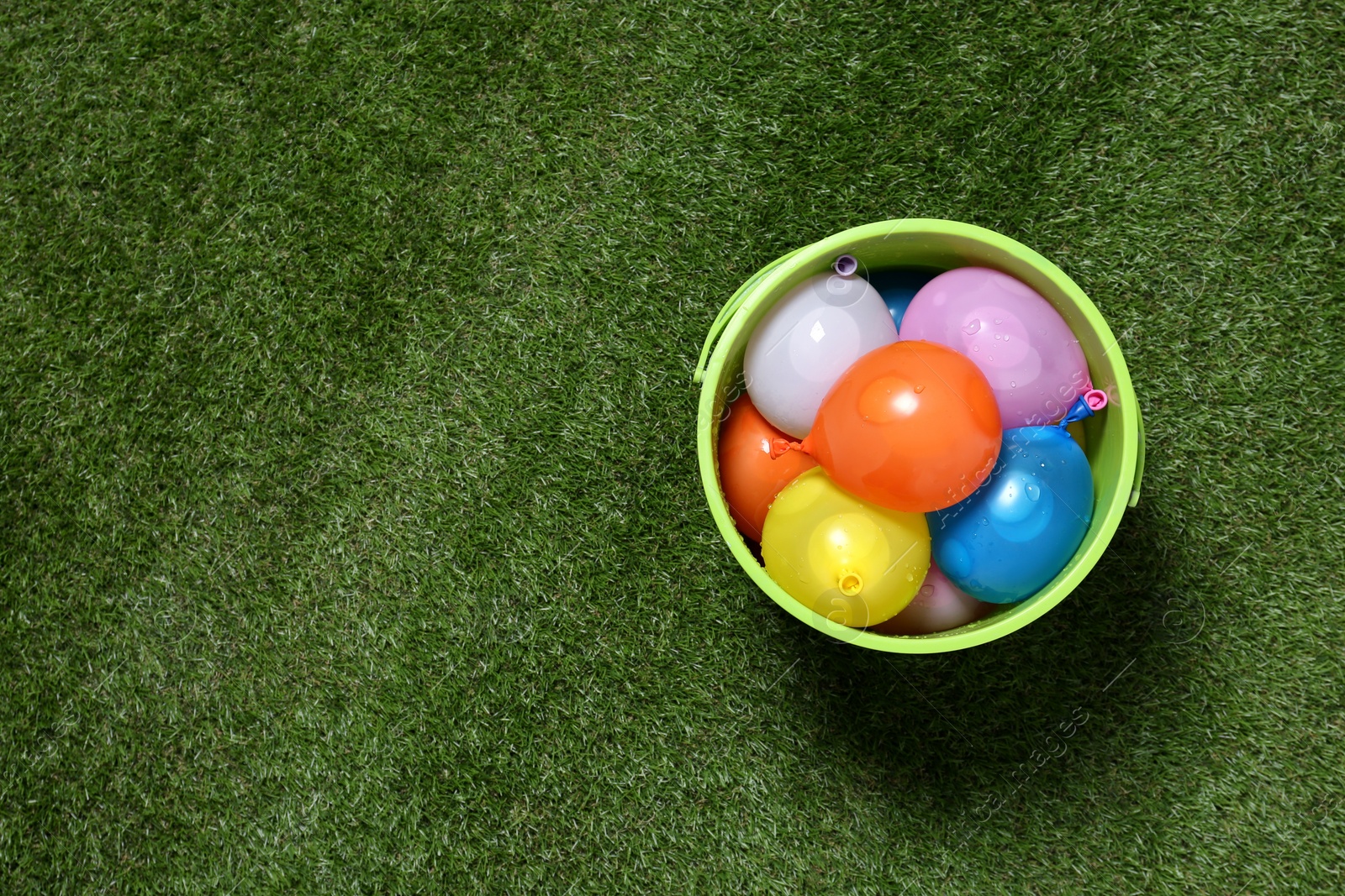 Photo of Bucket full of colorful water bombs on green grass, top view. Space for text