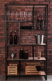 Rack with bottles of wine and glasses near brick wall