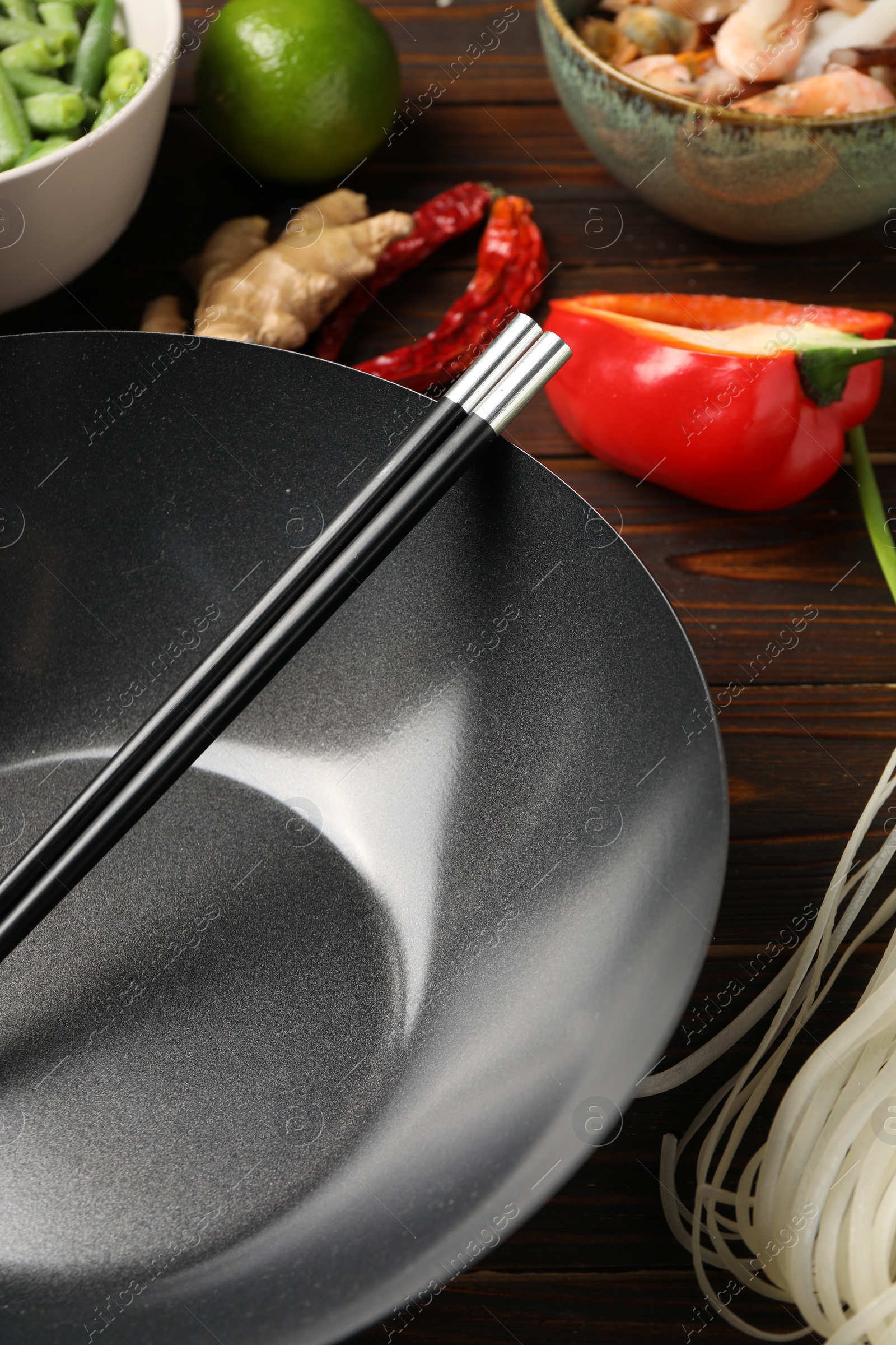 Photo of Black wok, chopsticks and products on wooden table, closeup