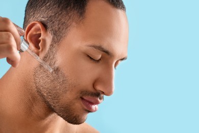 Handsome man applying cosmetic serum onto face on light blue background, closeup. Space for text