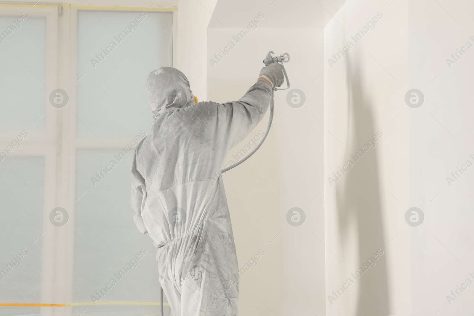 Photo of Decorator in protective overalls painting wall with spray gun indoors