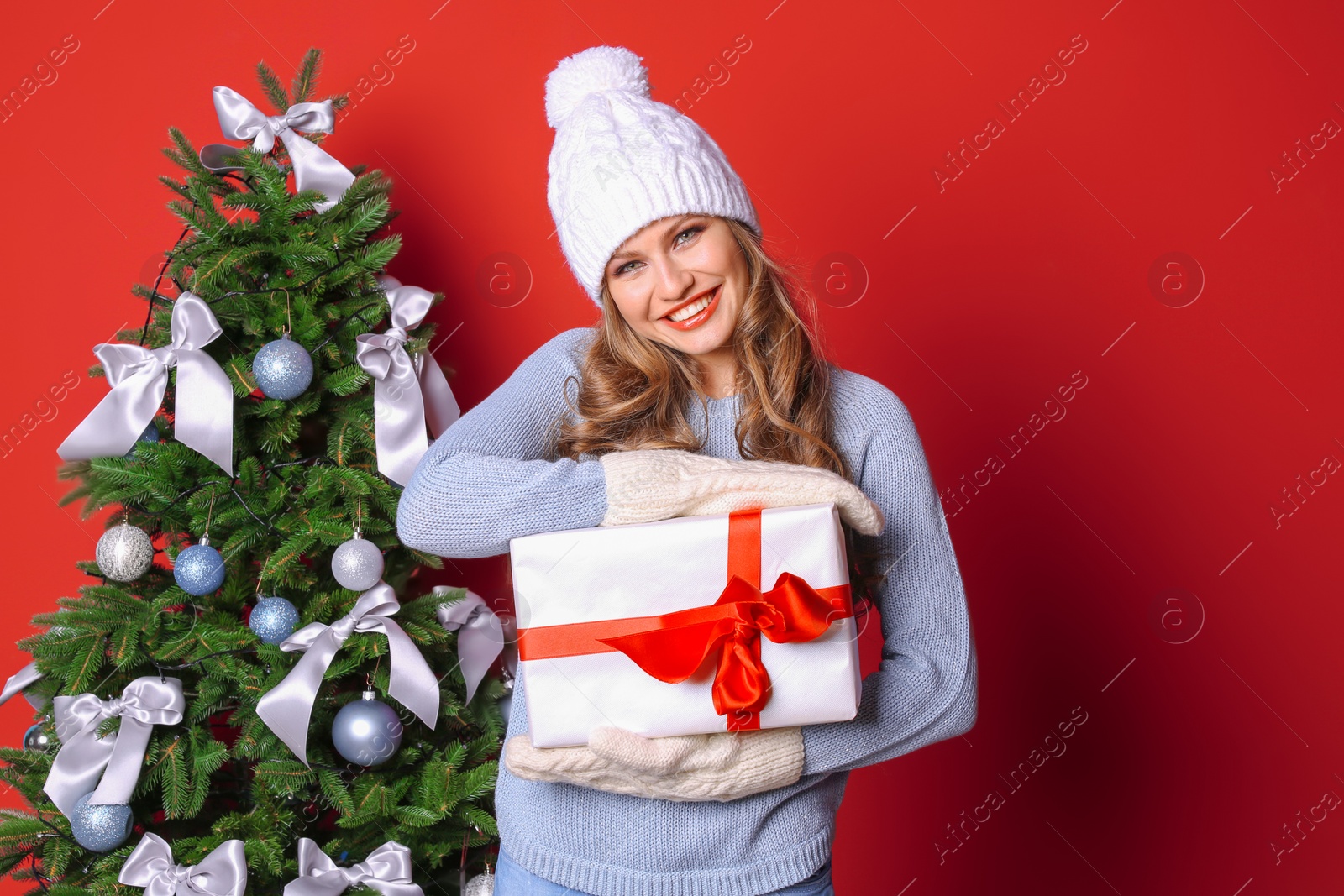 Photo of Beautiful young woman in hat and mittens with gift box near Christmas tree on color background