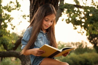 Photo of Cute little girl reading book near tree in park