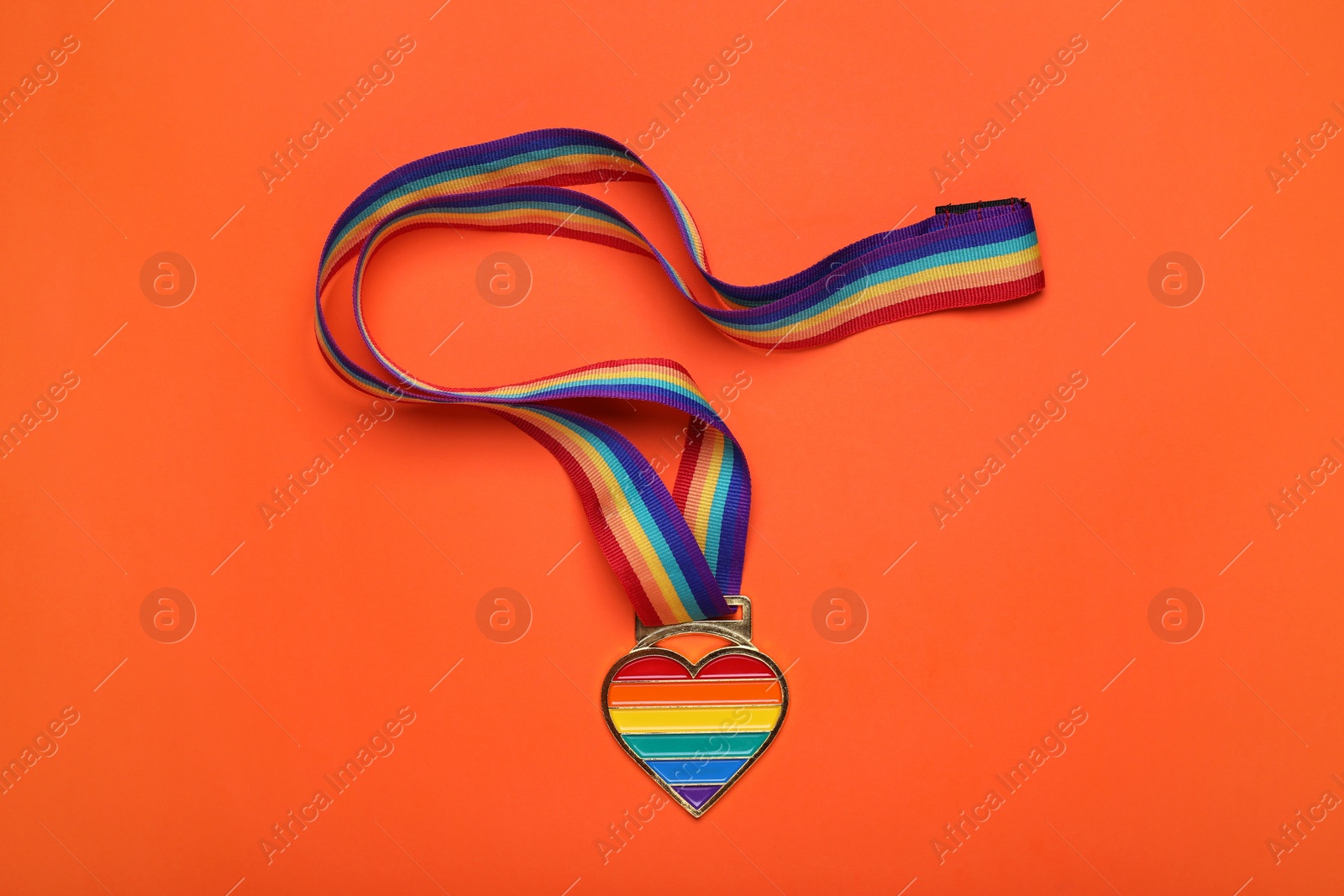 Photo of Rainbow ribbon with heart shaped pendant on orange background, top view. LGBT pride