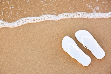 White flip flops on sand near sea, top view with space for text. Beach accessories