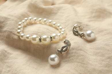 Photo of Elegant bracelet and silver earrings with pearls on beige fabric, closeup