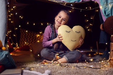 Happy girl with heart shaped pillow in decorated play tent at home