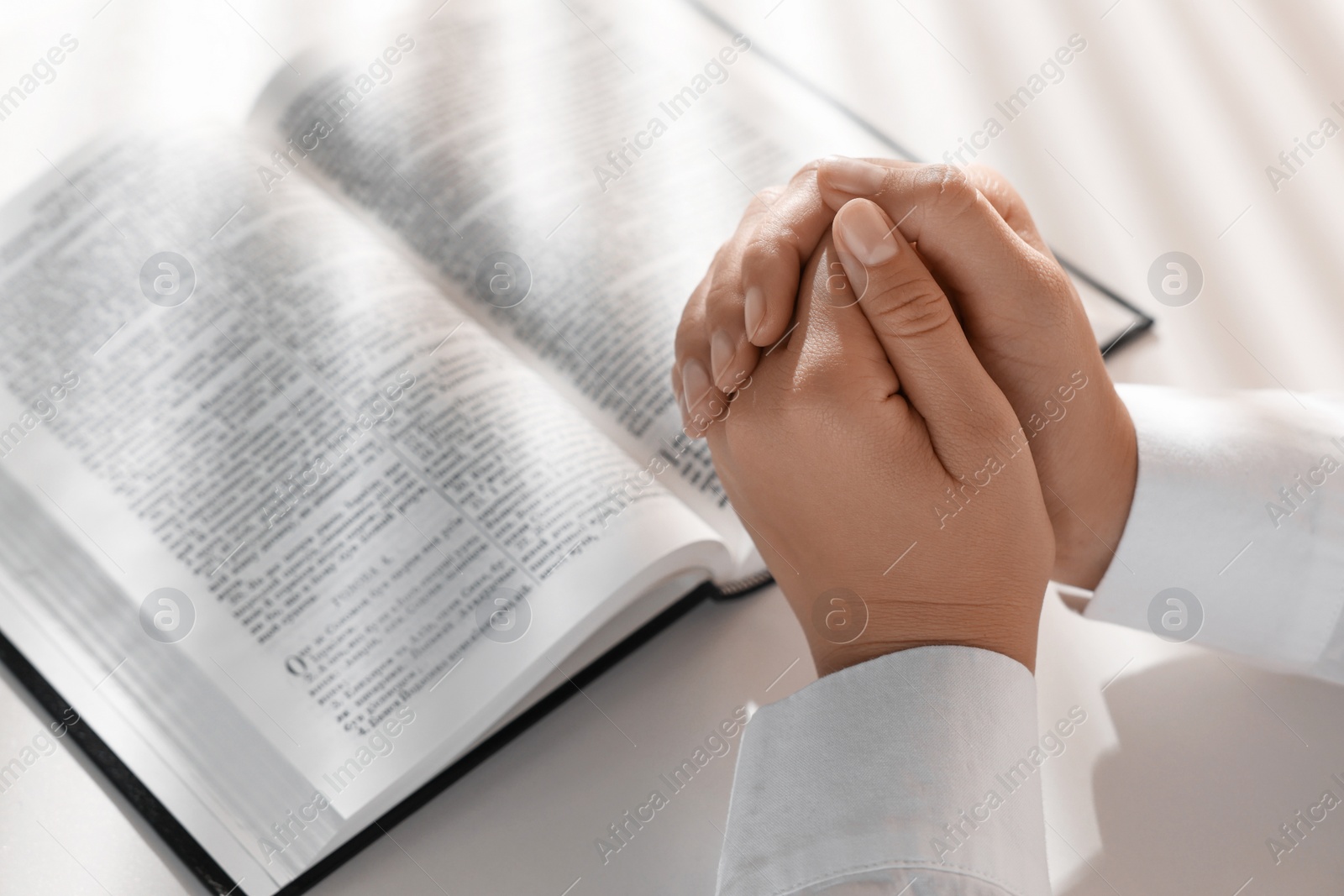 Photo of Woman holding hands clasped while praying over Bible at white table, closeup