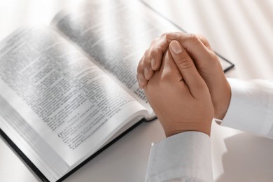Woman holding hands clasped while praying over Bible at white table, closeup