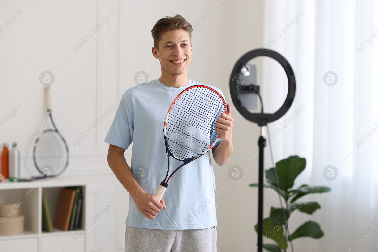 Photo of Smiling sports blogger holding tennis racket while streaming online fitness lesson at home