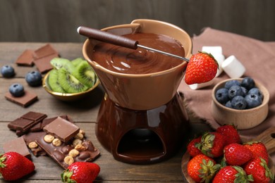 Photo of Fondue pot with melted chocolate, different fresh berries, kiwi and fork on wooden table