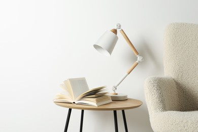 Photo of Stylish lamp with open book on wooden coffee table and soft armchair near white wall. Interior design