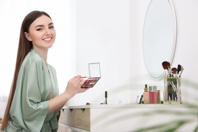Photo of Beautiful young woman with eyeshadow palette and brush at dressing table indoors