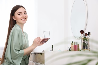 Beautiful young woman with eyeshadow palette and brush at dressing table indoors