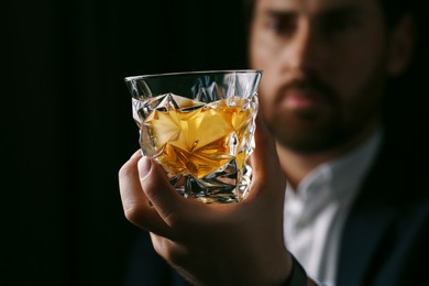 Man holding glass of whiskey with ice cubes on black background, selective focus