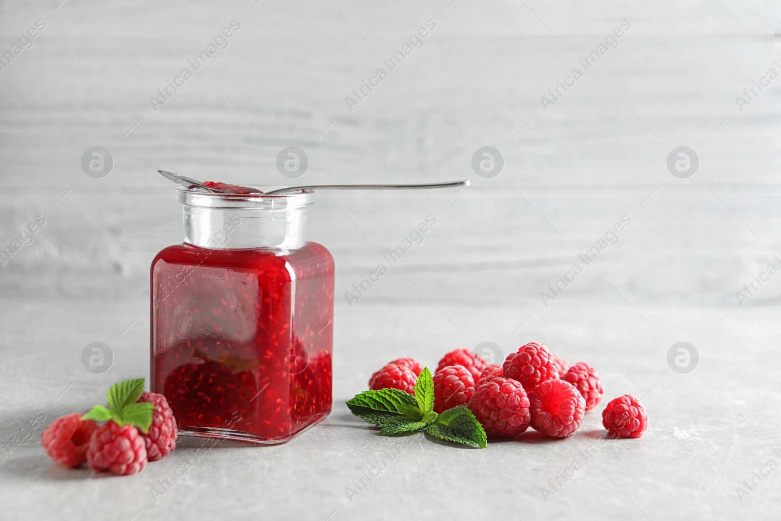 Photo of Glass jar and spoon of sweet jam with ripe raspberries on table. Space for text