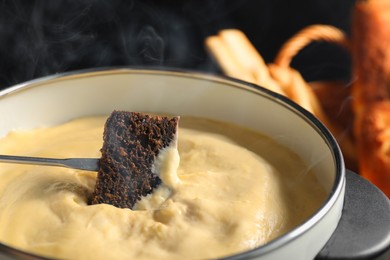 Photo of Dipping piece of bread into fondue pot with melted cheese on black background, closeup