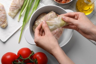 Woman putting uncooked stuffed cabbage roll into ceramic pot at white table, top view