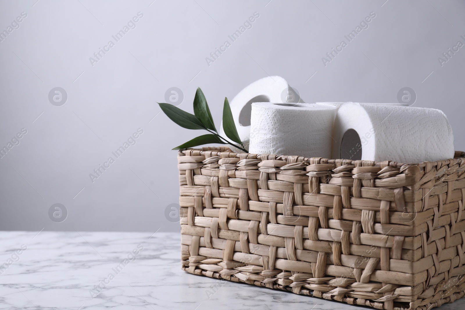 Photo of Toilet paper rolls and green leaves in wicker basket on white marble table, space for text