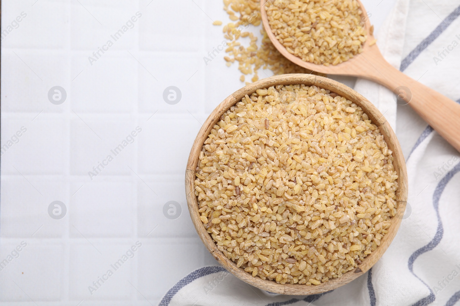 Photo of Bowl and spoon with raw bulgur on white tiled table, flat lay. Space for text