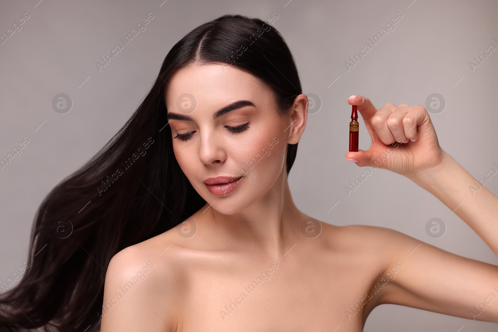 Photo of Beautiful young woman with long healthy hair holding ampoule on grey background