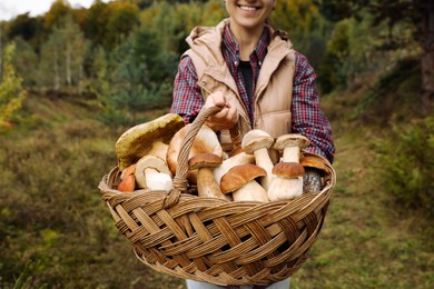 Photo of Woman holding wicker basket with fresh wild mushrooms outdoors
