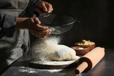 Woman sprinkling flour over dough at black table, closeup