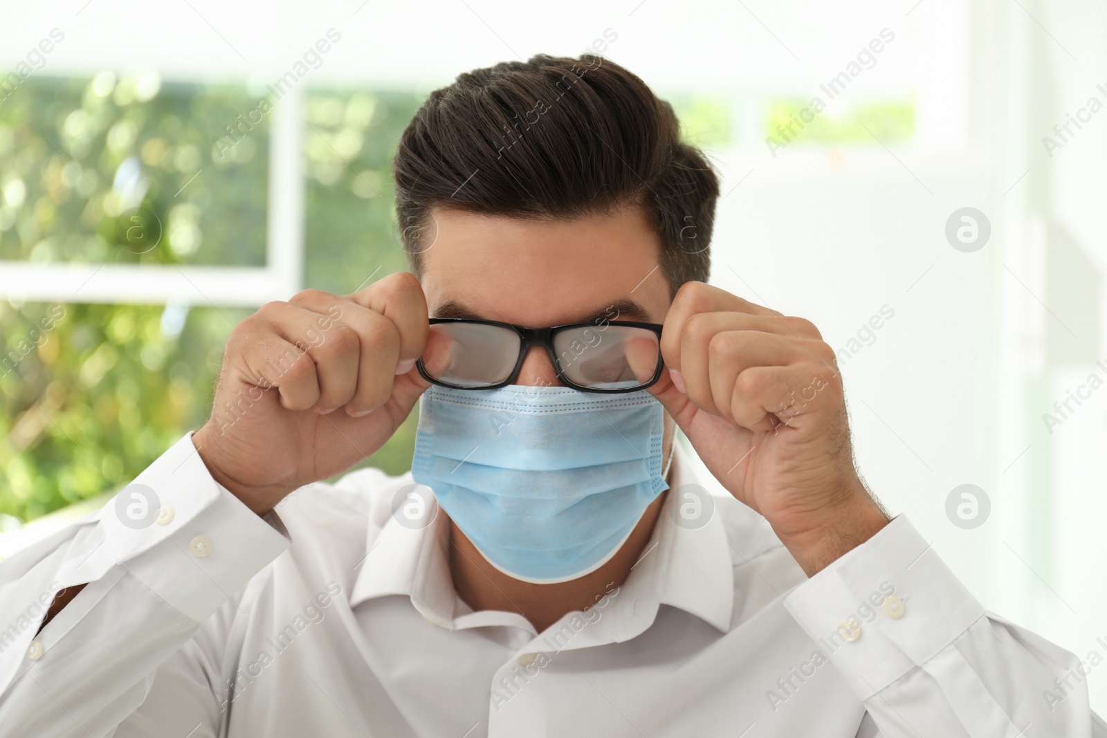 Photo of Man wiping foggy glasses caused by wearing medical mask indoors, closeup